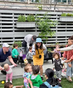 Karla in a yellow pinafore and blue bucket hat smiles while interacting with children outdoors, surrounded by parents and caregivers.